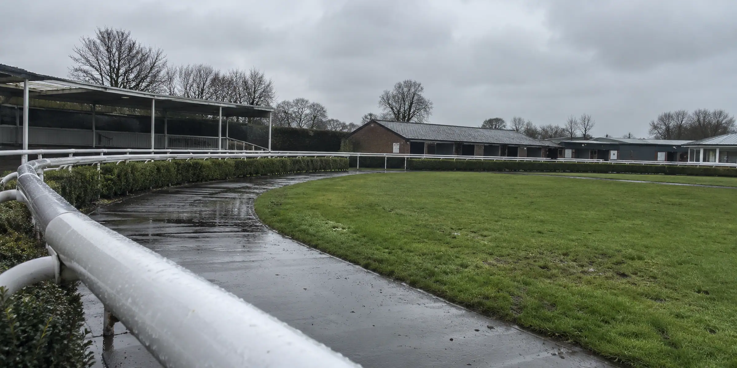 An empty parade ring with a wet path and white rails under grey skies